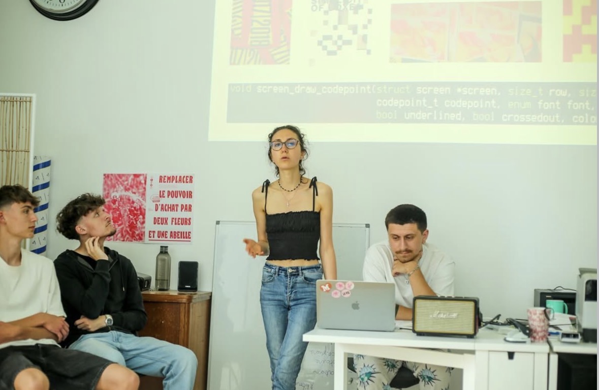 Maud Lenoir présente un projet debout devant un petit groupe assis, un écran affiche du code